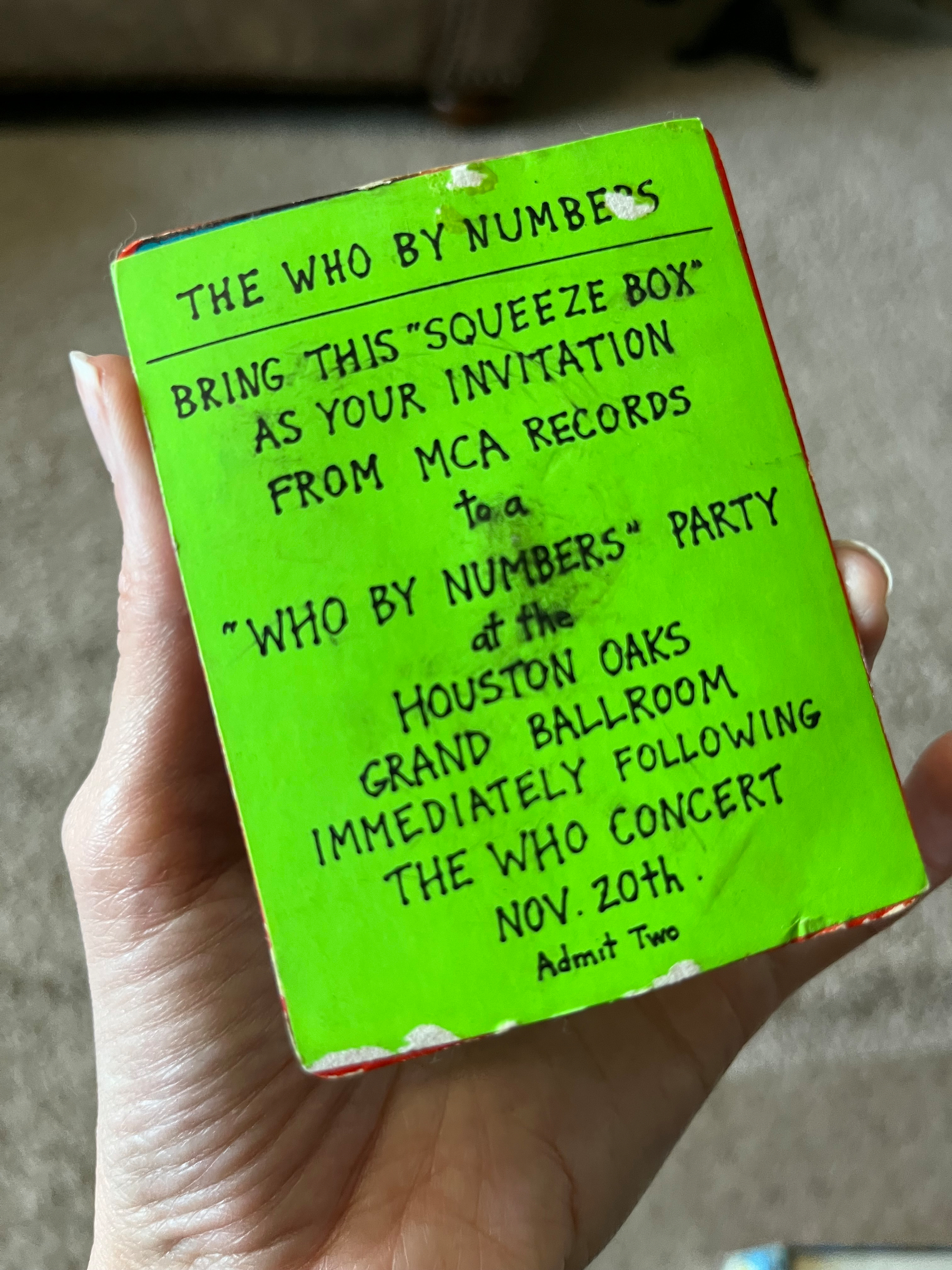 A hand holds an invitation to a Who By Numbers party in a bright green sleeve, associated with a concert in Houston.